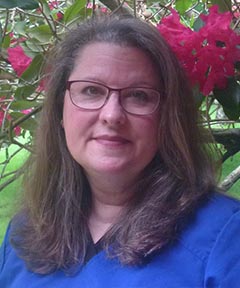 A woman with glasses, smiling slightly, poses against a backdrop of pink flowers and lush greenery.