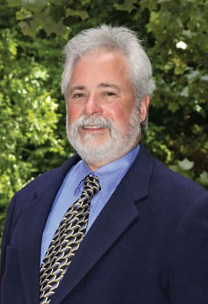 The image shows a man standing outdoors, wearing a suit with a tie and a beard. He has gray hair and is smiling at the camera.