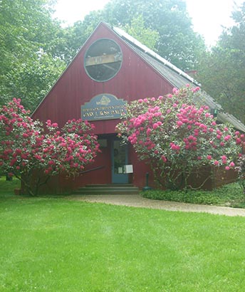 The image shows a red building with a sign that reads  The Old School House  surrounded by blooming pink flowers, set against a backdrop of a clear sky.