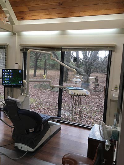 The image shows an interior view of a dental office with a modern chair for dental procedures, a computer monitor displaying patient information, and a large window allowing natural light into the room.