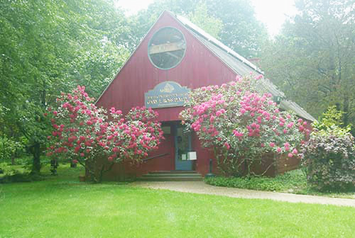 The image depicts a red building with a prominent sign, surrounded by lush greenery and blooming trees, on a cloudy day.