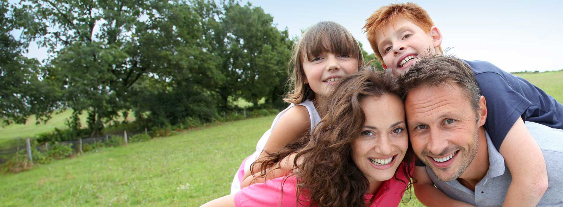 The image shows a family of four with two adults and two children posing for a photo outdoors during daylight, with a blurred landscape in the background.