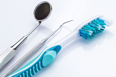 The image shows a collection of dental tools, including a toothbrush with blue bristles, a pair of scissors with silver handles, and a spoon-shaped instrument, all arranged on a white surface against a plain background.