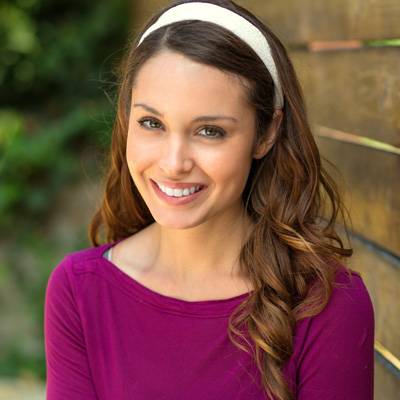 A young woman with long brown hair is smiling at the camera, wearing a purple top and a white headband. She stands against a wooden fence background.