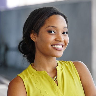 The image shows a woman smiling at the camera with her hair pulled back, wearing a yellow top and a necklace, standing against a blurred background that suggests an urban setting.
