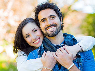 The image features a man and woman in a warm embrace, with the man wearing a scarf around his neck. They are outdoors, standing on a grassy area, and appear to be enjoying each other s company.