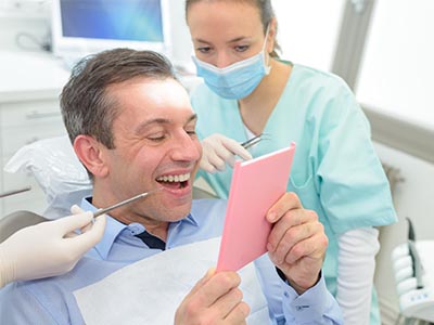 The image shows a man sitting in a dental chair, holding up a pink card with a surprised expression, while a woman in scrubs holds a mirror and smiles at him.