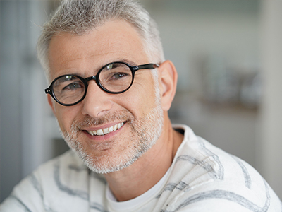 The image features a man with grey hair and glasses, smiling at the camera, wearing a white shirt with a patterned collar, set against a blurred background that suggests an indoor setting.