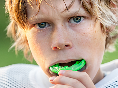 The image shows a young boy with blonde hair, wearing a white football jersey, holding a green toothbrush in his mouth while looking directly at the camera with a neutral expression.